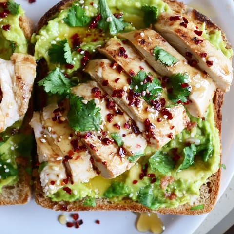 Golden-brown grilled chicken rests on vibrant green avocado mash over crusty toast with a honey-chili garnish.  